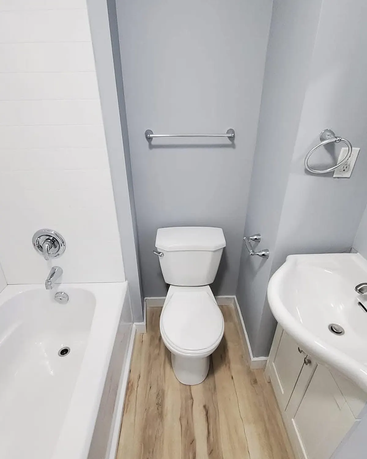A finished bathroom: a white tub with a tiled surround, a toilet, a pedestal sink on a white cabinet, and wood-look plank flooring.