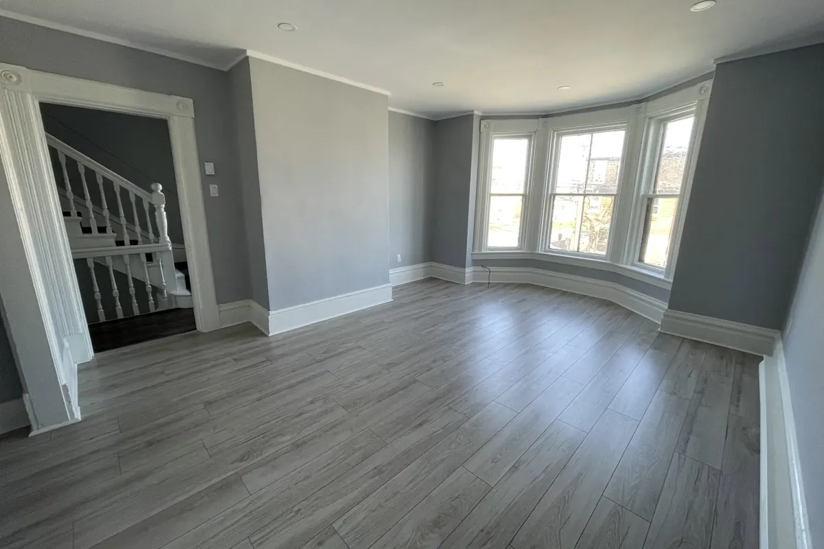 A long room with grey plank floors running to a bay window, a staircase visible through the doorway.