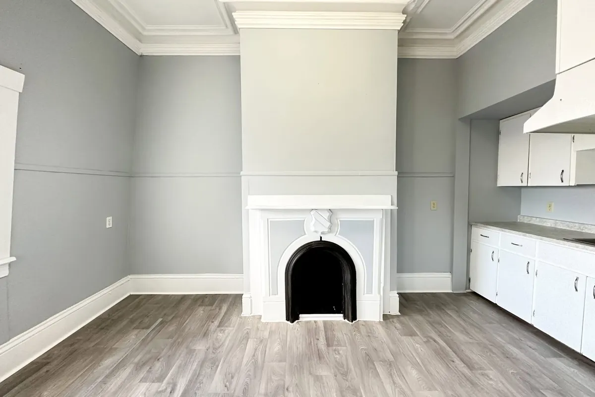 A room with grey plank floors, a white arched fireplace surround and white kitchen cabinets along the side wall.