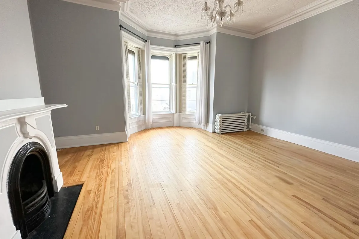A long living room with honey hardwood floors, grey walls, a white mantel around a cast-iron fireplace, a bay window and a chandelier.