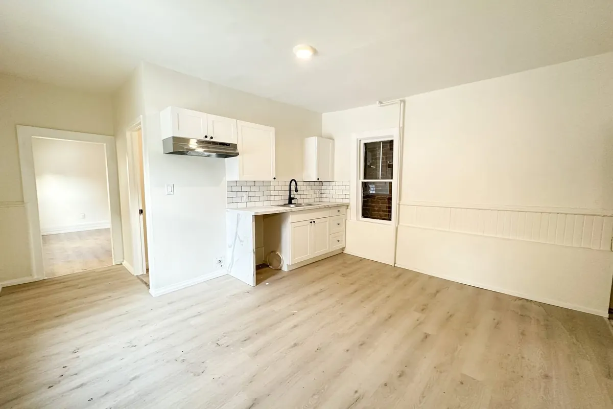 A kitchen with white cabinets and a subway-tile backsplash, open to a living room with light plank floors.