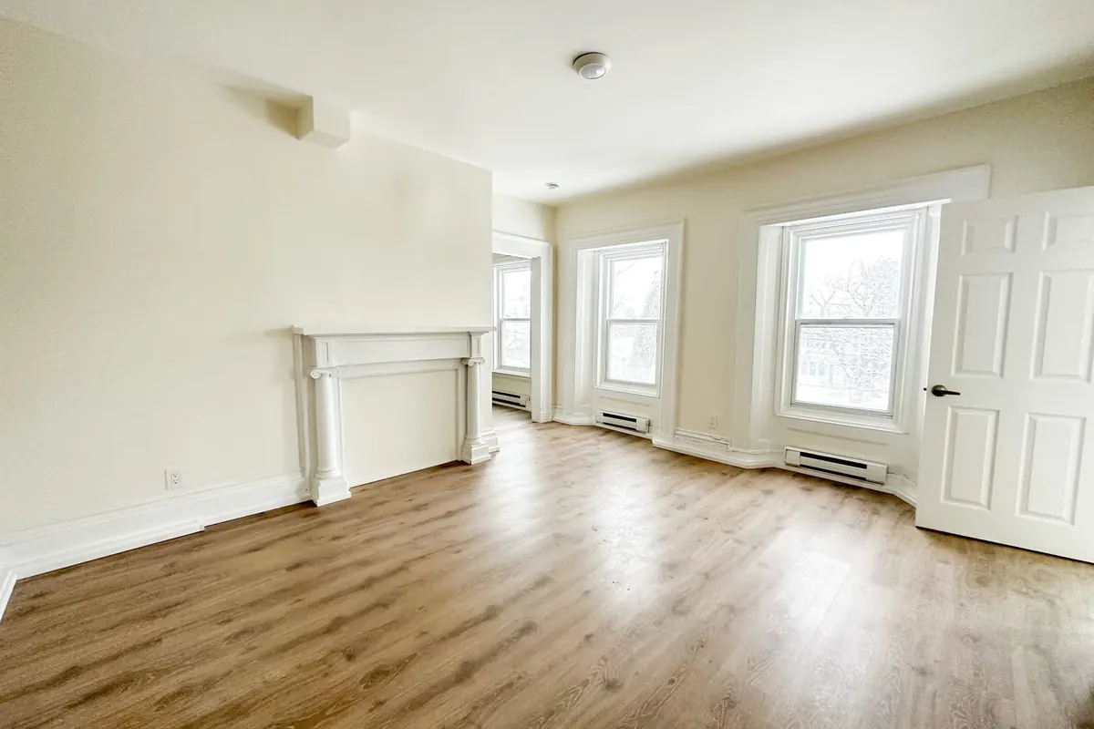 A bright front room with light plank floors, cream walls, a white mantel and tall windows.